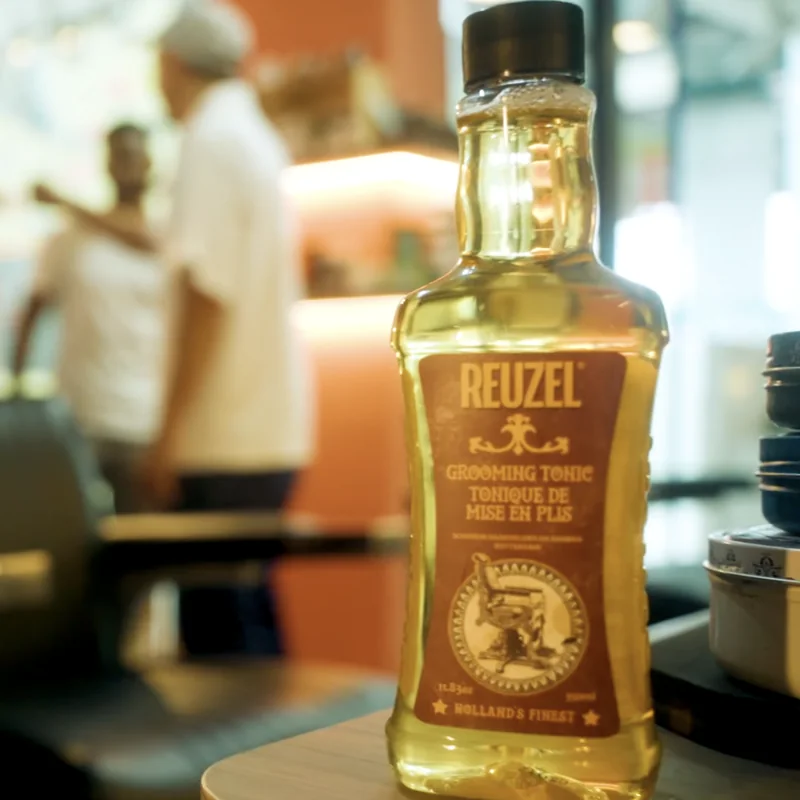 Reuzel Grooming Tonic bottle on a barbershop counter, with barbers blurred in the background